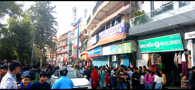 A busy street scene in Nahan, Himachal Pradesh, with students and people gathered outside shops, including the BRC Institute and Himachal Emporium. Vehicles, trees, and multi-story buildings are visible.
